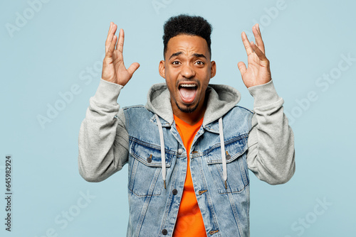 Bilde på lerret Young sad mad furious man of African American ethnicity wear denim jacket orange t-shirt spread hands scream look camera isolated on plain pastel light blue cyan background studio