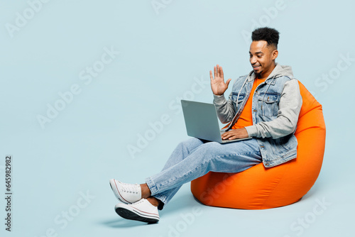 Full body young IT man of African American ethnicity wear denim jacket orange t-shirt sit in bag chair hold use work on laptop pc computer waving hand isolated on plain pastel light blue background.