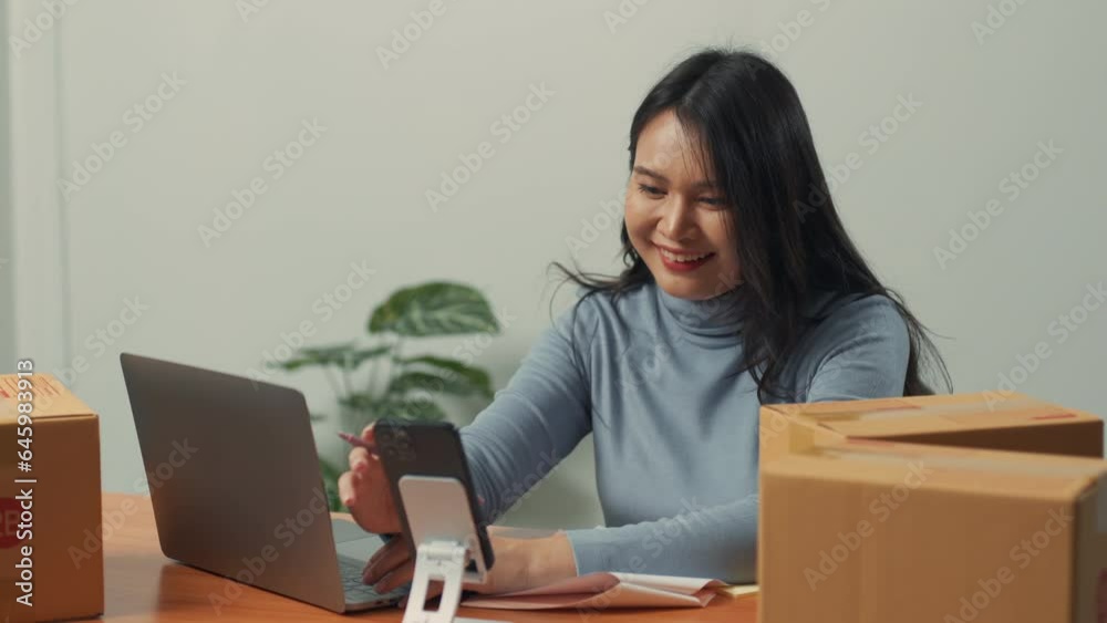 Asian woman checking sales on smartphone and working on laptop computer from home on table with parcel, e-commerce business, online sales concept Asian woman checking sales on smartphone and working on laptop computer from home on table with parcel, e-commerce business, online sales concept