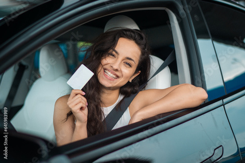 Cheerful young lady sitting in her car and showing her new driving license. Driving test, driver courses, exam concept