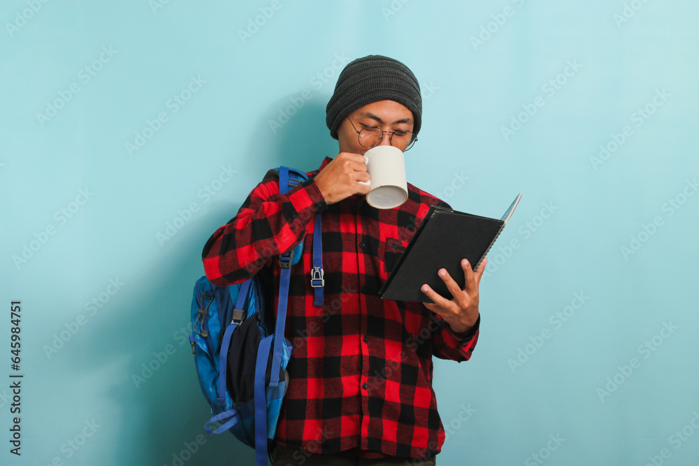 Young Asian male student takes a coffee break after lectures, isolated on blue background Young Asian male student takes a coffee break after lectures, isolated on blue background