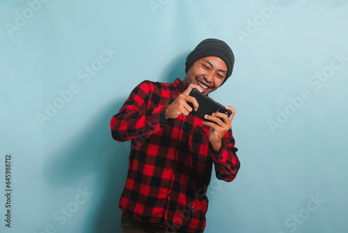 Excited young Asian man with a beanie hat and a red plaid flannel shirt is playing a racing game on his smartphone, isolated on a blue background