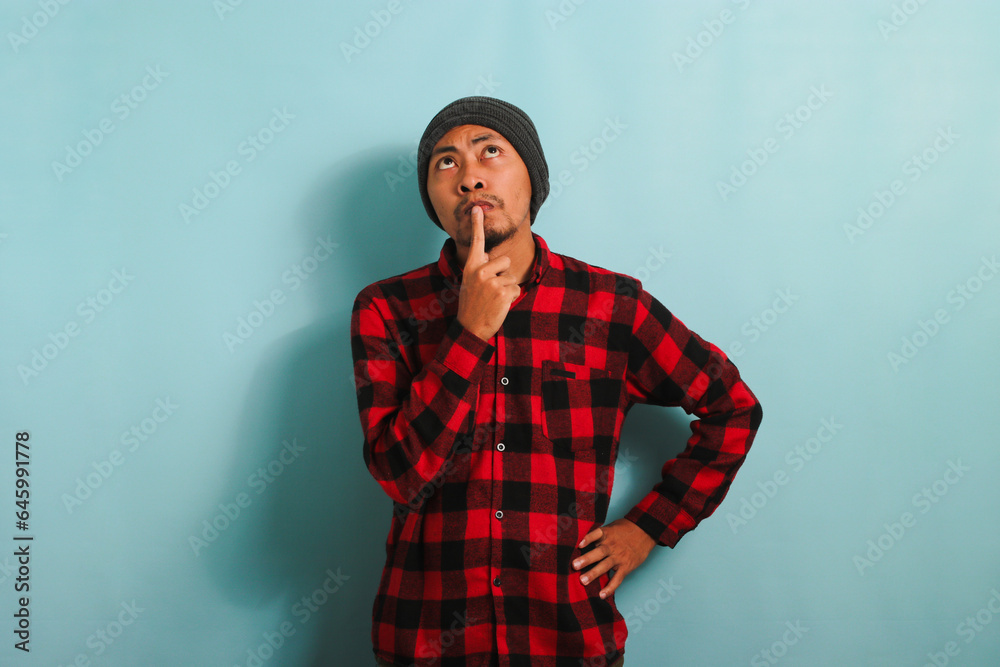 Pensive young Asian man keeps his hand on his chin, looking at copy space, isolated on blue backdrop