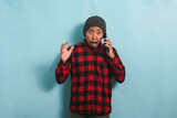 Shocked Young Asian man with a beanie hat and a red plaid flannel shirt is surprised and raises his hand while talking on his mobile phone, isolated on a blue background