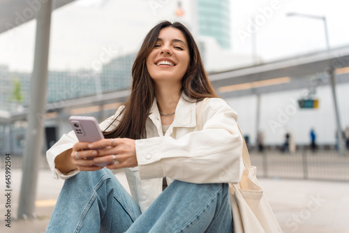 Waiting for the bus and for love. Bright cheerful young woman of Turkish mixed race in white casual clothes with a charming smile holding a phone in the street at public transportation stop