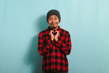 Smiling young Asian man with a beanie hat and a red plaid flannel shirt shows a love gesture with his hand while standing against a blue background