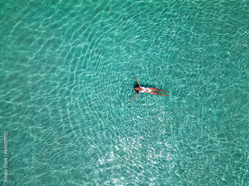 Top view of a young woman in the transparent, blue ocean. Aerial view of a swimming woman lying and floating on the water in the Maldives