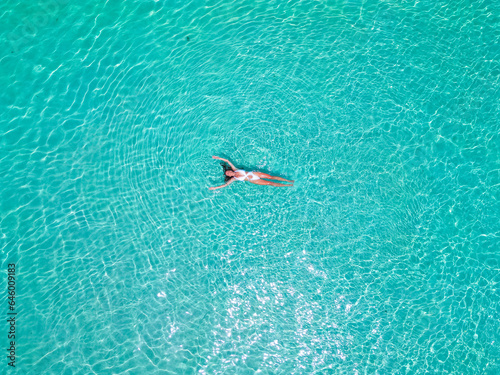 Top aerial drone view of a girl in a white swimsuit relaxing, swimming, and sunbathing lying on the sparkling clear blu water in the ocean in the Maldives