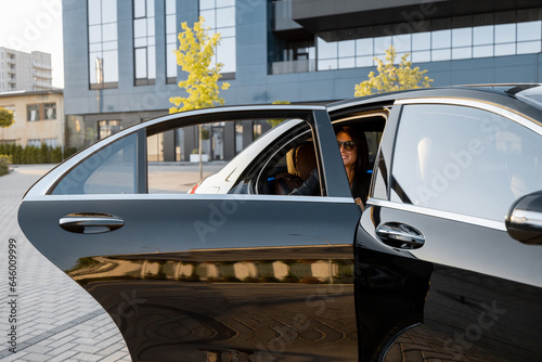 Elegant business woman gets in luxury black car near office building or hotel. Concept of business transportation and travel