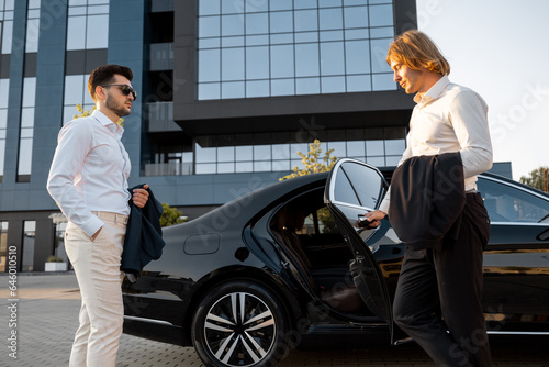 Two businessmen get in luxury car near hotel or office building on sunset, going to drive after work. Concept of transportation and business lifestyle