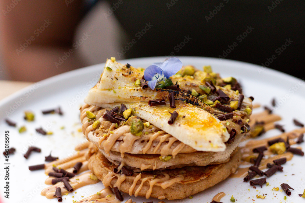 A stack of banana pancakes with slices of fresh bananas, pistachios and honey on top with chocolate slices and edible flower. Close up