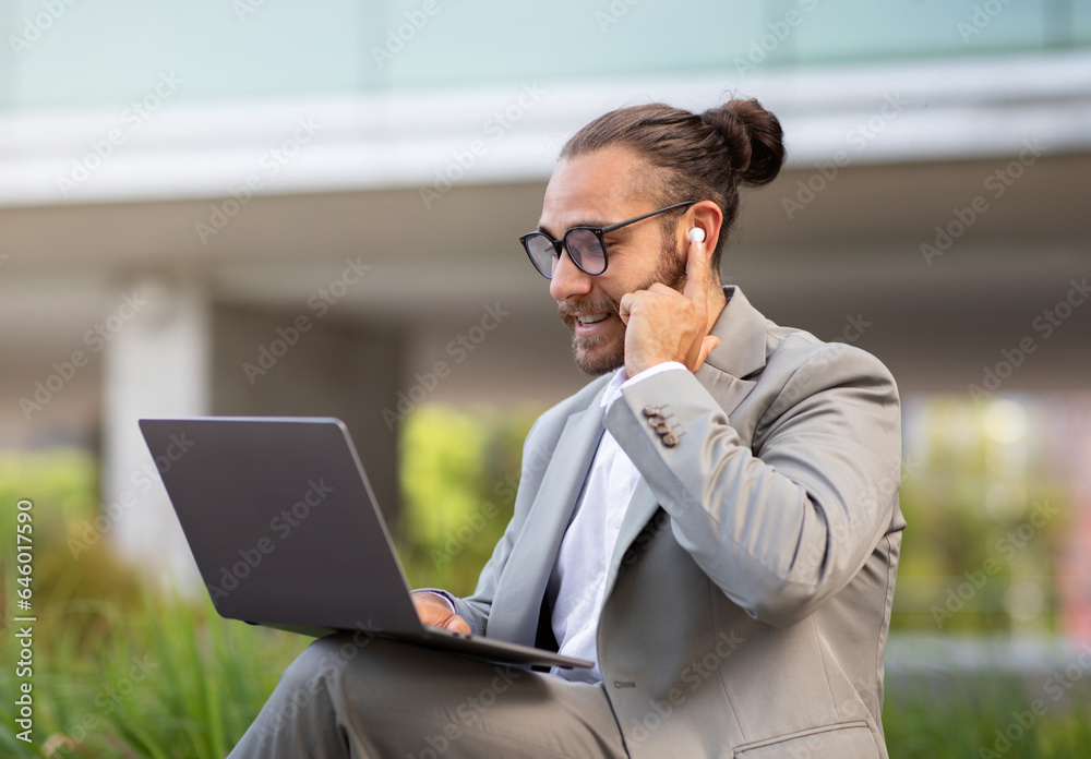 Handsome millennial businessman making video call on laptop while sitting outdoors Handsome millennial businessman making video call on laptop while sitting outdoors
