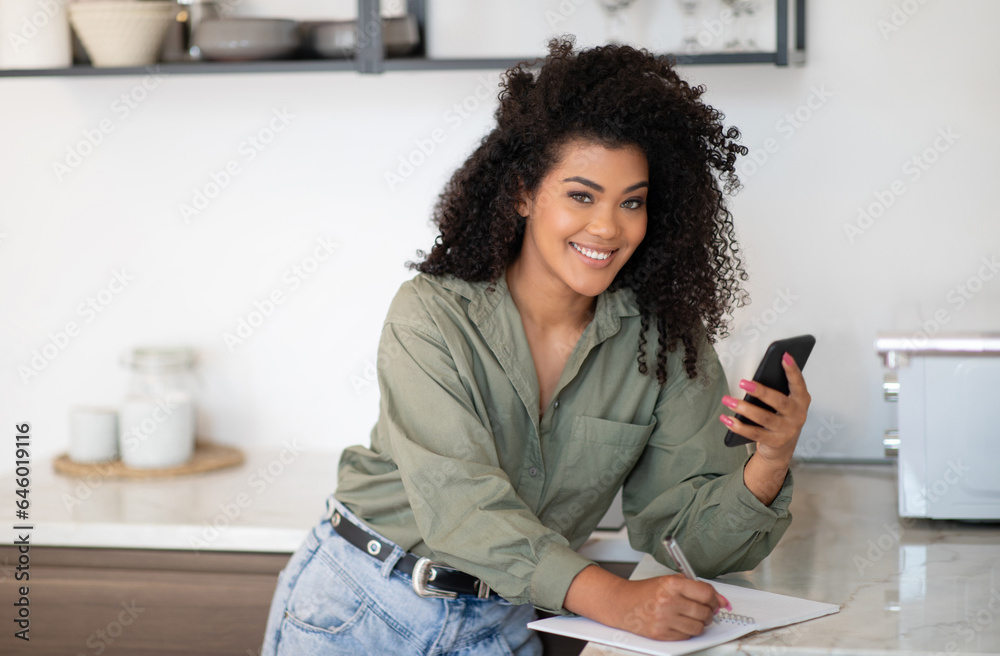 Smiling African American Lady Using Cellphone Taking Notes In Kitchen