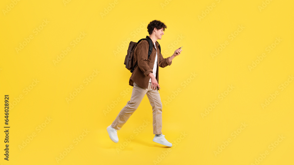 Fototapeta premium Cheerful Student Guy Using Smartphone Walking With Backpack, Yellow Background