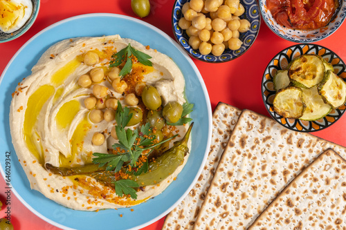 Hummus plate, matzah bread, chickpeas, baked vegetables on red background.
