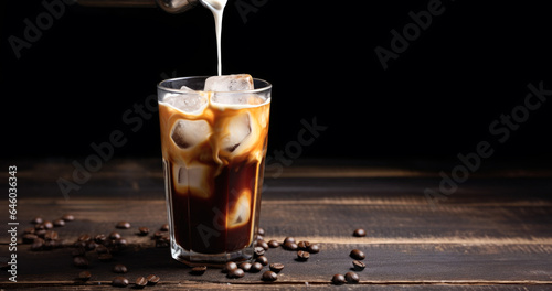 milk pouring into glass of ice coffee or cold brew on a wooden table next to roasted coffee beans.