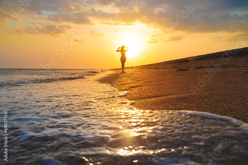 Seascape during golden sunrise or sunset with beautiful sky. Woman on the beach. Young happy woman in a straw hat walks along the seashore. Girl looks at the magical sunrise