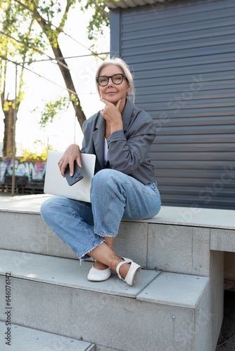 charming stylish mature woman with glasses dressed in a gray jacket works remotely sitting with a laptop on the street