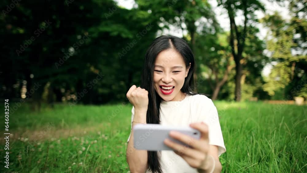 Happy excited young asian woman gamer sitting in the park and playing in online video games on smartphone holding in horizontal landscape mode outdoors Happy excited young asian woman gamer sitting in the park and playing in online video games on smartphone holding in horizontal landscape mode outdoors