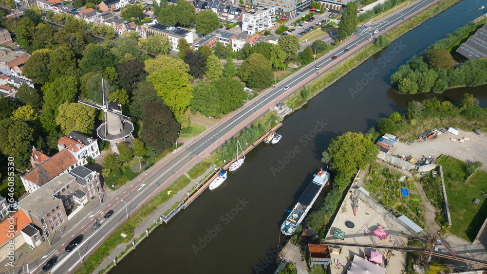 Aerial drone shot of treetops, trees, houses and shop rooftops of Dutch city Gouda centre with historic building het slot windmill with nieuwe veerstal road and Hollandsche Ijssel on clear sunny day