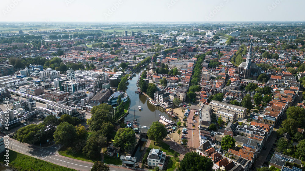 Fototapeta premium Aerial drone shot of treetops, trees, houses and shop rooftops of Dutch city Gouda centre with historic building het Tolhuis or toll house with nieuwe veerstal road on clear sunny day