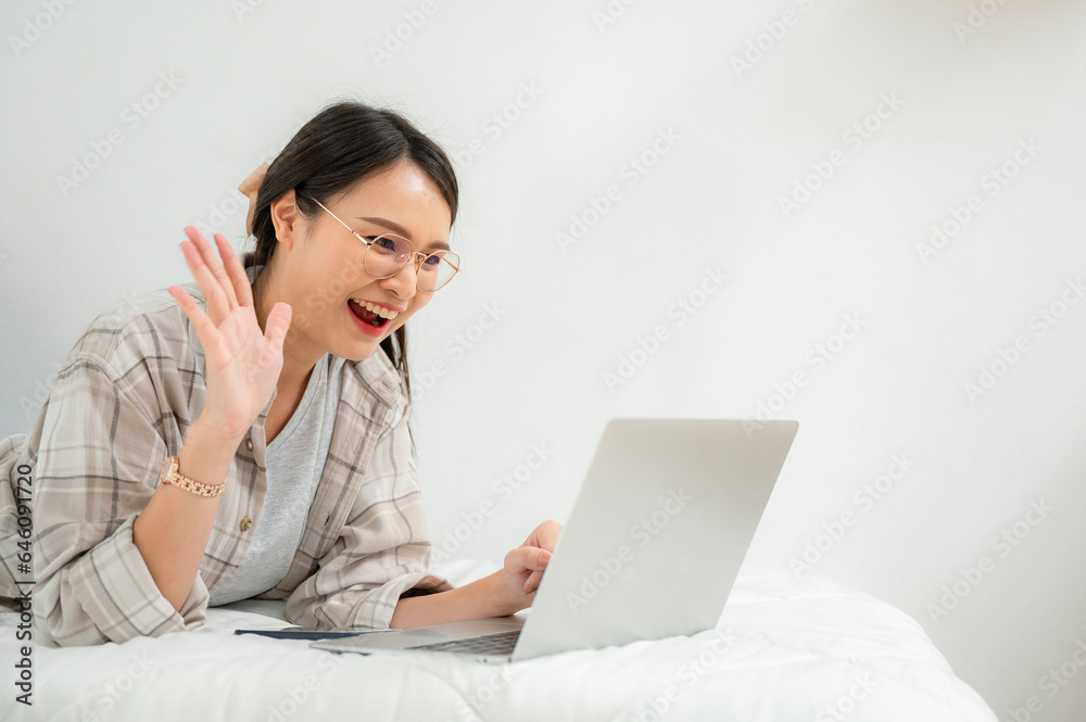 Happy Asian young woman freelancer talking online remotely on laptop at her bed at home. Happy Asian young woman freelancer talking online remotely on laptop at her bed at home.