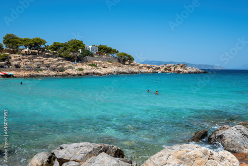 Greece Aponisos beach Agistri island. Rocky beach pine tree umbrella, people swim in turquoise sea