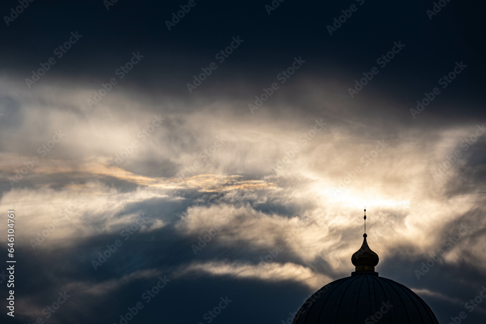 the dark mysterious profile of the round dome of an Orthodox church with a thin cross at the top against the background of a beautiful sunset sky with feathery clouds where the rays of the evening sun