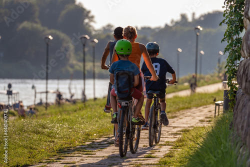A happy family rides bicycles along the river bank on a summer day towards the sun. Lifestyle and travel concept. Rear view of a group of bikers of different ages wearing helmets. Close-up