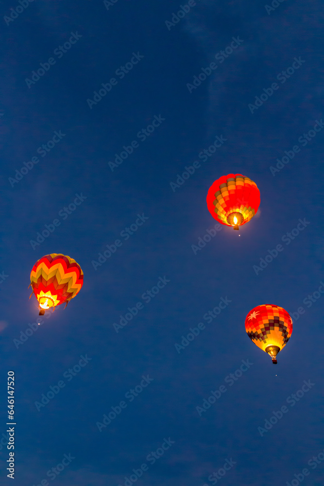 Fototapeta premium 3 hot air balloons are rising in the early morning light into a dark blue and wispy clouded sky. The 3 balloons are brightly lit with their gas burners on, The are brightly colored.