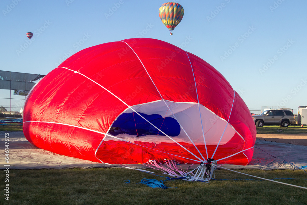 Obraz premium A red hot air balloon is on the ground being deflated. Ropes are coming from the entrance of the balloon. The interior is white and blue. 2 hot air balloons are in the background.