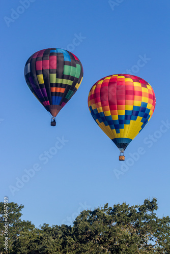 2 hot air balloons are rising in the early morning light into a blue and wispy clouded sky, 1 balloon is slightly above the balloons, The are brightly colored.