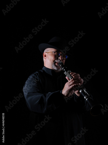 Senior adult with white beard and round glasses making music on music day with handmade Egyptian flute decorated with inlay in front of black piano with hat. Black background. Low key
