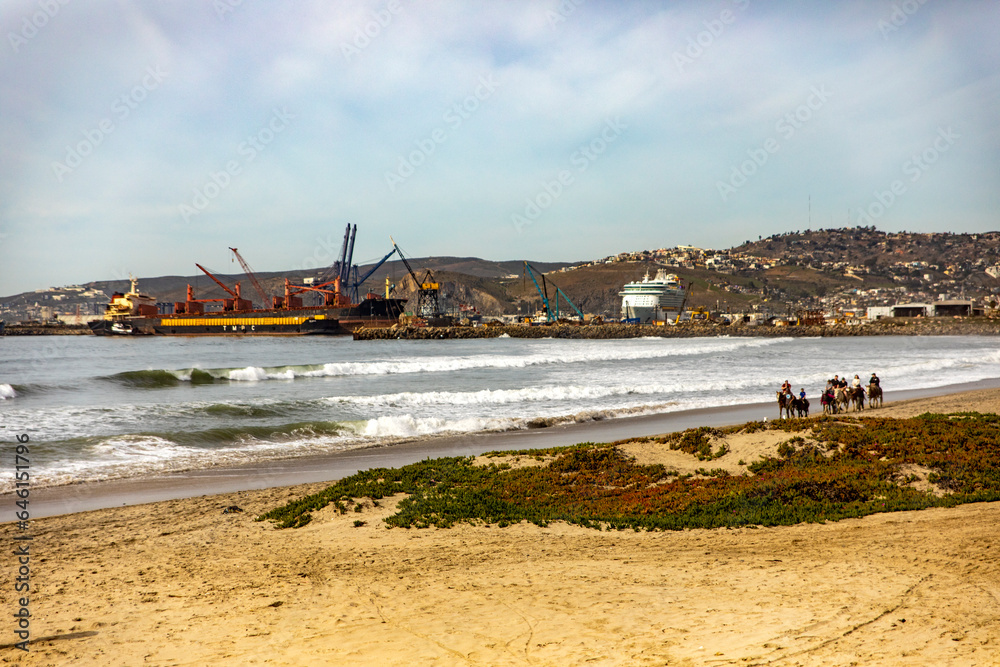 A beautiful beach in Ensenada with the port in the background and people riding horses by the sea, this is one of the best tourist places in Baja California, Mexico.