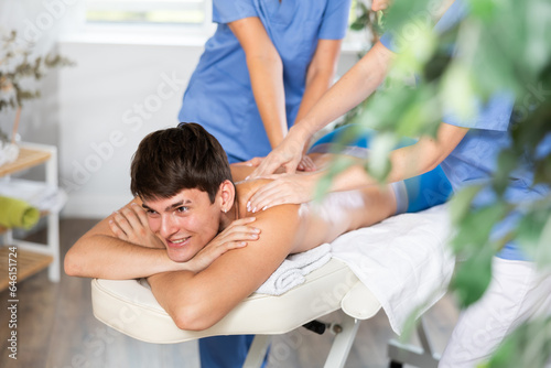 Massage in four hands. Two unrecognizable female masseuses perform stroking and rubbing of muscles of back and shoulders to young guy client of spa salon © JackF