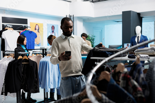 African american man shopping for outfit in clothing store and checking size options in smartphone app. Mall client searching for garment information on mobile phone website