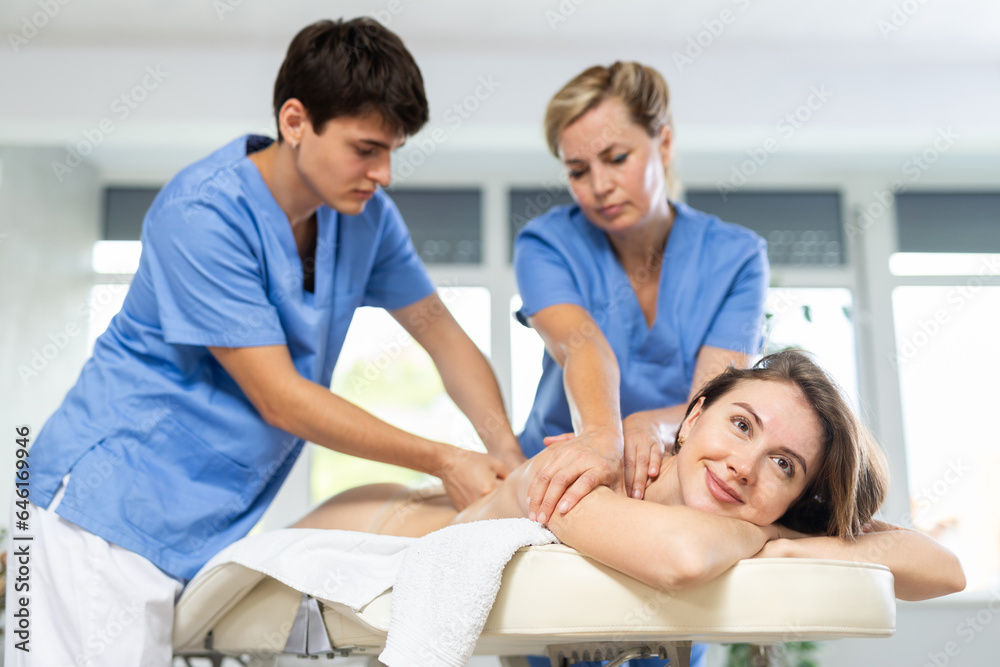 Relaxed young woman undergoing body massage conducted by two masseurs in therapy cabinet