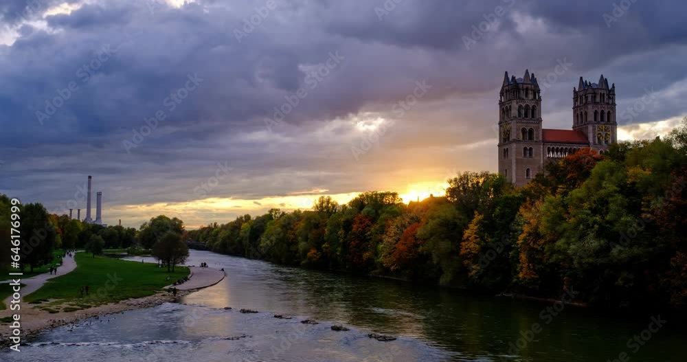 Timelapse of Munich at dramatic sunset: autumn in famous tourist landmark Bavarian city. View of Isar river, trees, and church from Reichenbach Bridge. Munchen, Bavaria, Germany. Camera zoom in