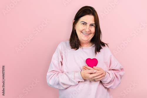 A mature woman smiles while holding a pink resin heart, symbolizing hope and support for breast cancer awareness, ideal for October Pink campaigns © Marcio