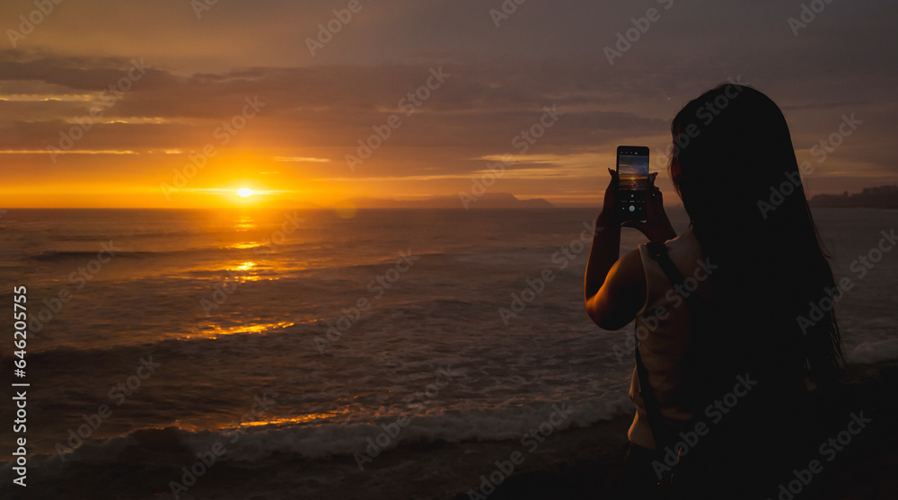 Young woman backlit against the sea at sunset using the phone. Space for text.
