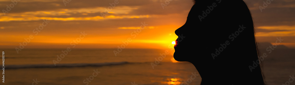 Silhouette of a girl backlit, kissing the sun in front of the sea at sunset. Space for text.
