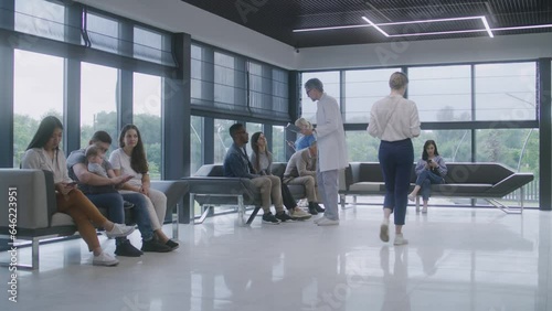 Multicultural people sit on sofas in clinic lobby area, wait for doctors appointment. Doctor talks to patient about medical test results. Waiting room in modern medical center. Healthcare. Dolly shot.