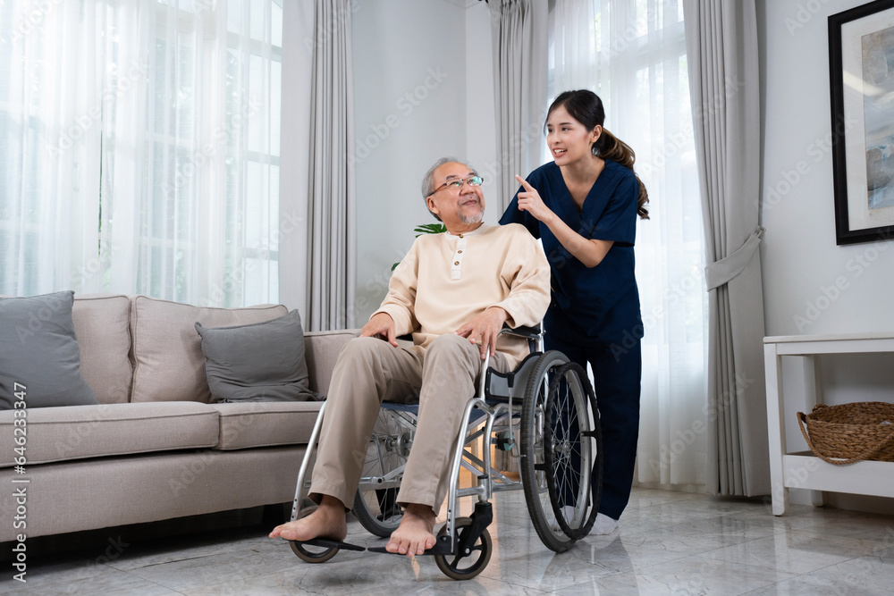 A young Asian female nurse or physical therapist in a blue uniform, standing and talking happily behind a senior Asian man sitting on a wheelchair while pushing him to relax in the house living room. A young Asian female nurse or physical therapist in a blue uniform, standing and talking happily behind a senior Asian man sitting on a wheelchair while pushing him to relax in the house living room.