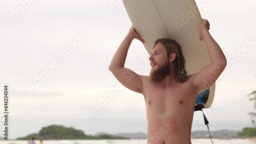 4K Portrait of Young Caucasian man surfer in swimwear holding surfboard walking on the beach