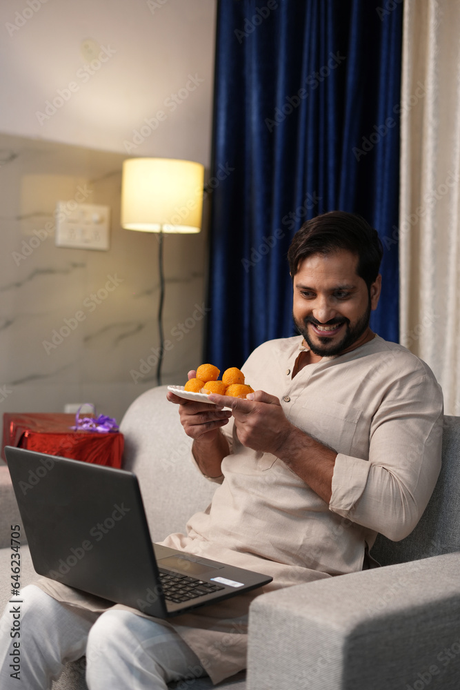 Young Indian man sitting on sofa holding plate of laddoo and using laptop