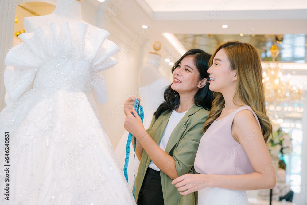 Asian bride is smiling while choosing wedding dress in modern wedding salon,  Attractive designer girl using tape meter fitting on wedding dress at wedding studio