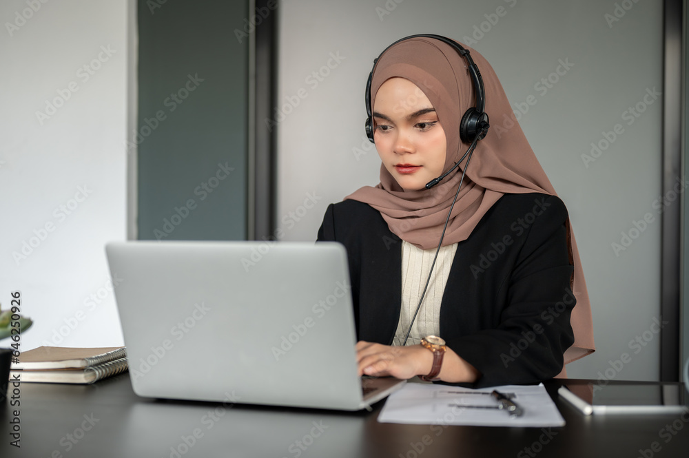 A focused Asian Muslim female call centre operator wearing a headset is answering an incoming call. A focused Asian Muslim female call centre operator wearing a headset is answering an incoming call.