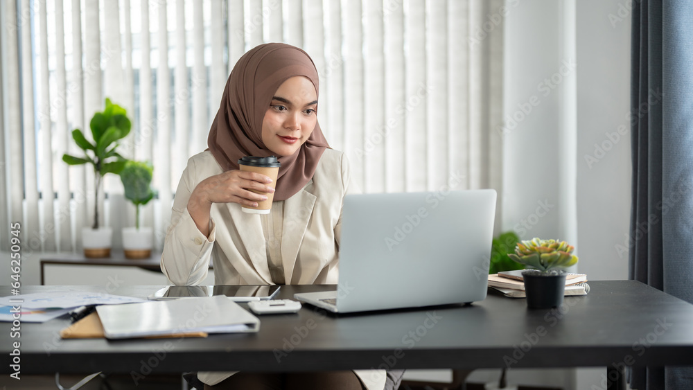 A focused Asian Muslim businesswoman is having morning coffee while working on her laptop. A focused Asian Muslim businesswoman is having morning coffee while working on her laptop.