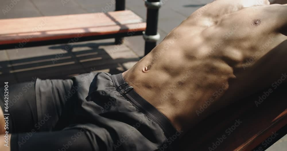 Closeup on shirtless man with athletic body doing reverse crunch exercise for abs muscles on bench at outdoors gym. Cropped view on young male with six pack has workout and abdominal training