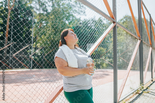 A fit, plus size woman stands outside and enjoys a moment with her water bottle after her exercise. Her dedication to a healthy lifestyle is unwavering. side view